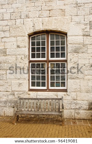 An old wooden bench on a sidewalk under a window in a stone block wall