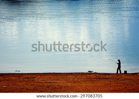 A man stands in front of the river to angle the fish with yellow sky reflecting on the water