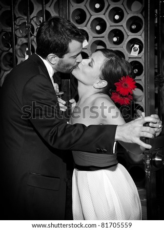 A bride and groom kissing in the wine cellar after their wedding. Black and white with a color treatment to bring out the red flowers in the bride\'s hair.