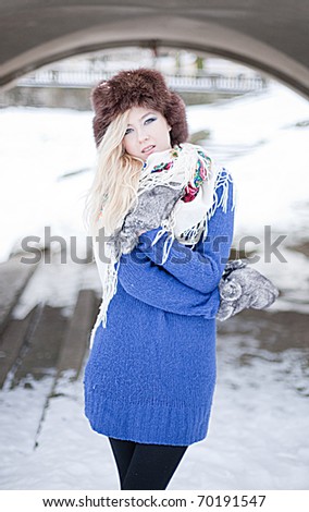Young caucasian blonde woman wearing blue sweater, furry hat with gloves and traditional shawl in winter scenery.