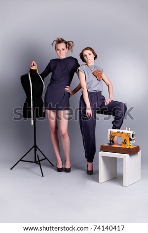 Fashion shoot: two young, sensual women in fashionable clothes with sewing mannequin and sewing machine on grey background