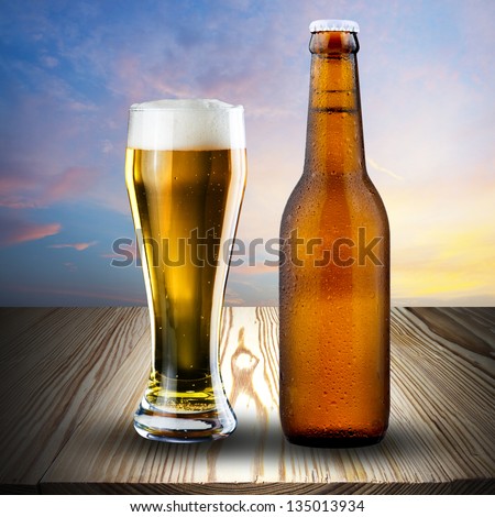 Bottle and glass of cold beer on wood with sunset sky background
