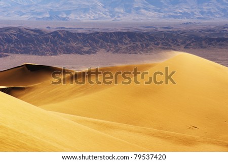 Strong wind blows the sand of the sand dunes in the desert with mountain in the background