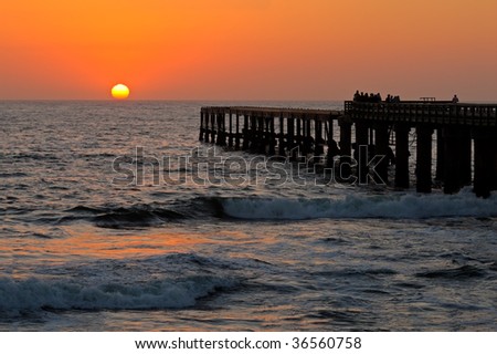 View of a silhouetted coastal pier with people watching a glowing sunset