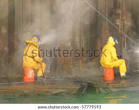 2 people washing an oily dock in hazard suits while sitting on bright orange buckets