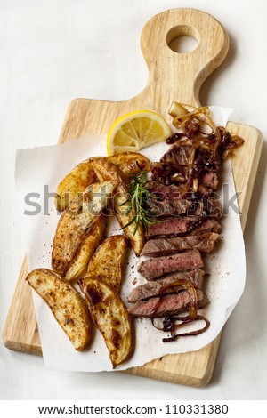 Sliced beef steak with potato wedges, onions, rosemary and lemon, on a board.
