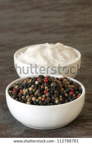 Black peppercorns and sea salt flakes in small white bowls, over stone background.