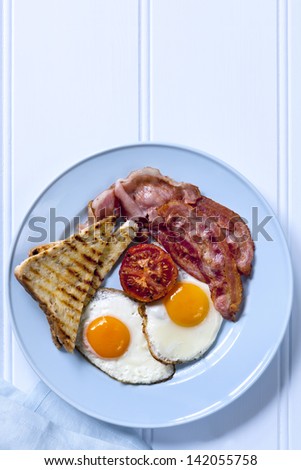 Bacon and eggs with tomato and toast.  Overhead view of blue plate on timber background.