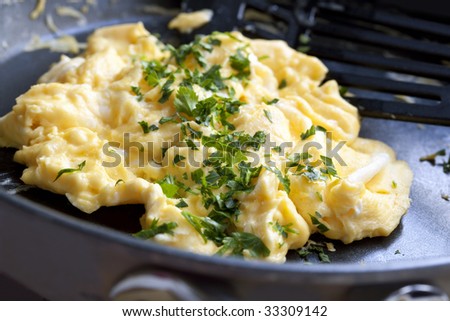 Scrambled eggs cooking in a frypan, with spatula.  Topped with flat-leaf parsley.