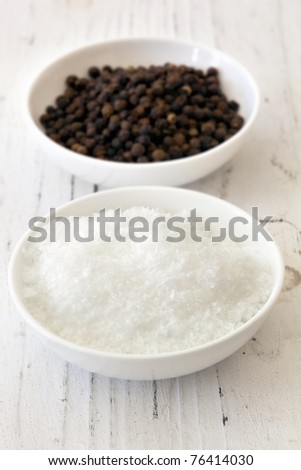 Sea salt flakes and black peppercorns, in small white dishes over weathered timber.