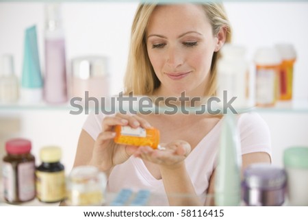 A young woman pours out medicine into her hand.  She is visible through her medicine cabinet.  Horizontal shot.