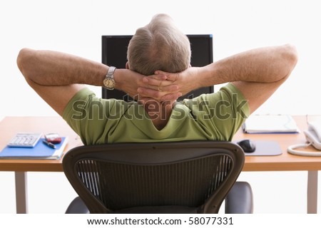Rear view of a man seated at a desk in front of a laptop. He is sitting back in his chair with his hands behind his head. Horizontal shot.
