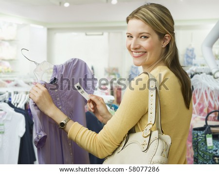 A young woman holds onto the price tag of a blouse while shopping.  She is smiling over her shoulder towards the camera.  Horizontal shot.