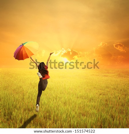 Red woman holding multicolored umbrella in green grass field and sunset