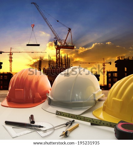 engineer working table plan, home model and writing tool equipment against building construction crane with evening dusky sky