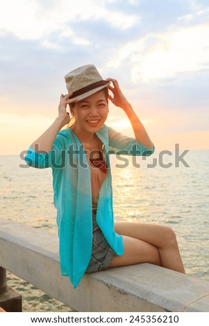portrait of beautiful and happy emotion teenager wearing straw hat sitting on bridge beside sea beach against sun set sky