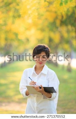 portrait of beautiful asian young woman standing in yellow flowers blooming park with writing tool for note some project ,check list something to do