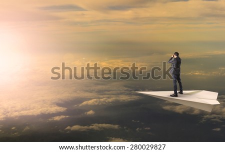 business man standing and spying binocular on paper plane against sun rising over cloud scape