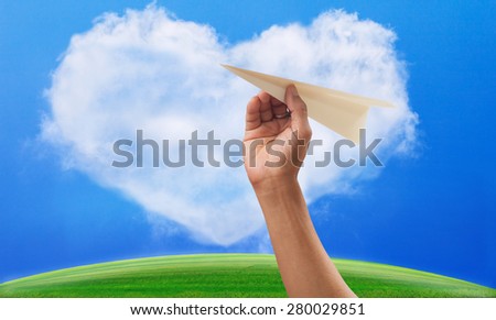 hand preparing to throwing paper plane to mid air against green grass field and heart shape white clouds on clear blue sky