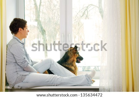 The girl with a short hairstyle in jeans clothes waits for someone sitting on a window sill and stroking a dog.