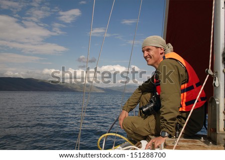 The smiling man in a life jacket sits on a nose of the yacht floating on the Lake Sevan Armenia