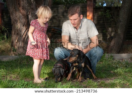 The father in a light shirt on a lawn shows to the fair-haired daughter of two quiet dogs