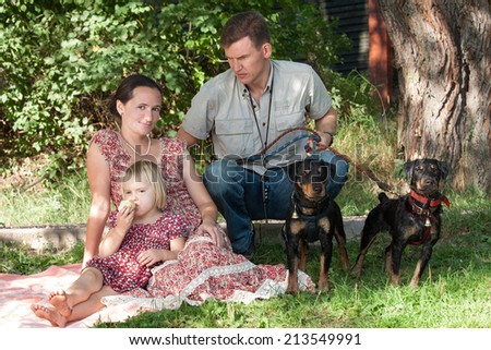 The young woman with the fair-haired daughter who eats apple, sit on a grass. The man who is sitting next and holding on a lead of two dogs looks at them.