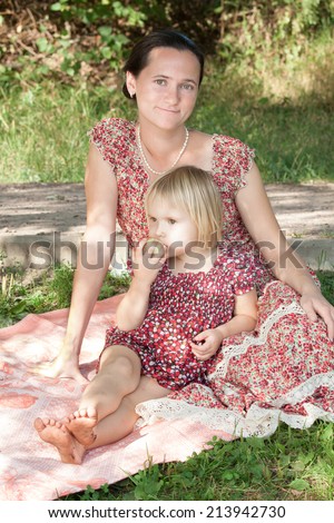 Mother with the daughter in summer dresses sit on a grass in park, the daughter relies on mother and, thoughtfully looking aside, gnaws apple