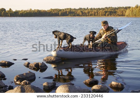 The man in a camouflage in the boat with two hunting dogs - Jagdterriers moors to the coast. Dogs watch in different directions, behind the back of the hunter a rifle, in hands an oar.