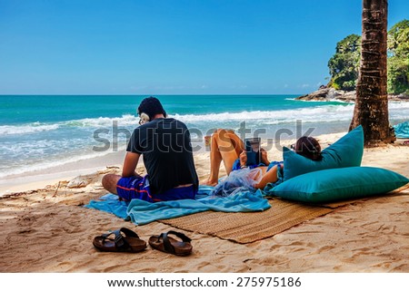 PHUKET, THAILAND- OCTOBER11, 2014: man listening music, woman using tablet on Surin beach. Surin is exclusive beach retreat for discerning travelers and couples seeking relaxation in a serene setting