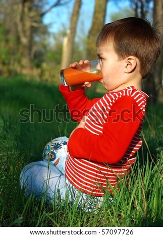 Boy drinks apple-carrot juice from transparent glass, sitting on grass. One hand keeps for stomach./Fourth glass of juice ...