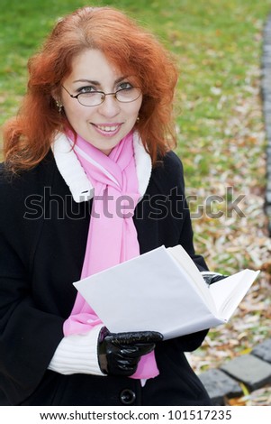 Young smiling red haired lady wearing glasses holding a book. The girl looks like a teacher or a student.