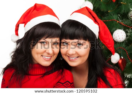 Two young dark-haired women in Santa\'s red caps smile near Christmas tree