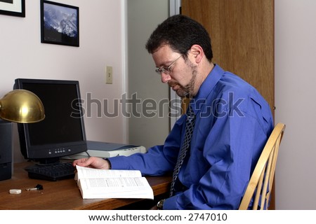 Middle-aged man reading a book in front of the computer in a home office.