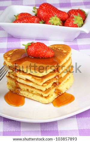 Stack of heart shaped pancakes with syrup and a strawberry on a white dish with fork. A perfect breakfast for Valentine\'s Day.
