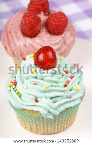 Two fancy cupcakes on a white porcelain plate. One with candied cherry and one with raspberries. Selective focus.