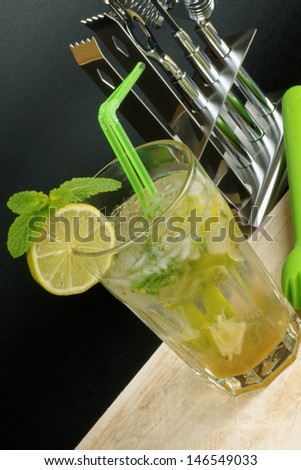 Mojito cocktail with two straws, lime slice and spearmint leaves and a cocktail set behind.