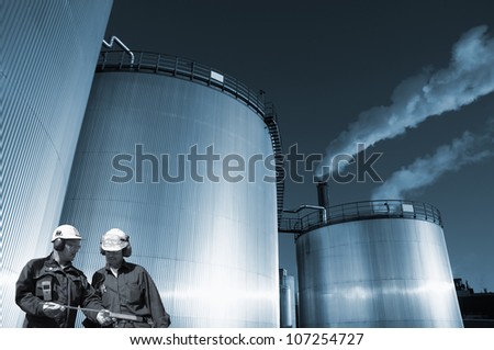 two oil and gas workers, large fuel storage tanks in background, blue toning concept