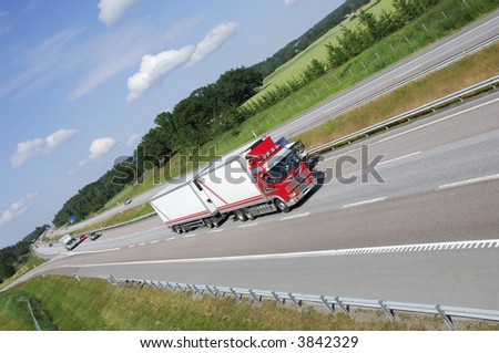 red and white truck speeding on highway with fields and forrest in background