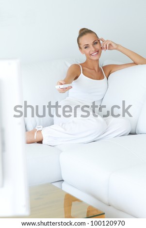 Pretty woman in casual white outfit sitting on a white couch with the remote control in her hand changing channels as she watches television