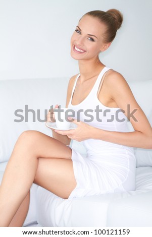 Glamorous woman with a beautiful smile sitting cross legged on a white sofa drinking coffee