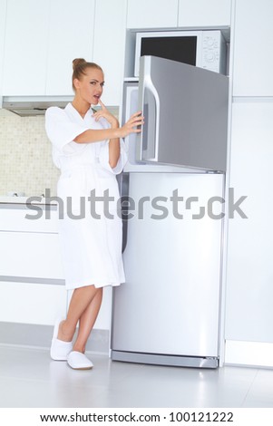 Vivacious woman in a white bathrobe and slippers standing laughing as she opens the freezer unit of her refrigerator