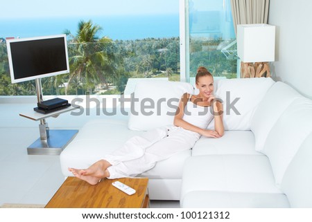 A beautiful woman relaxes on a white sofa in a modern airy bright living-room enjoying the luxury of a tropical lifestyle