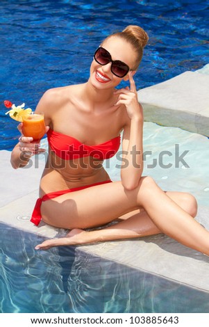 Pretty seated blonde with a beautiful smile wearing a small red bikini and holding a drink