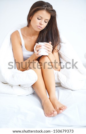 Woman drinking coffee in bed sitting in her pyjamas holding the mug cupped in her hands with a thoughtful downcast expression