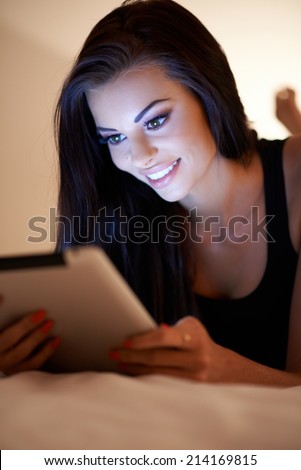 Young woman using her tablet computer at night smiling as she reads information on the screen with her face lit by the glow
