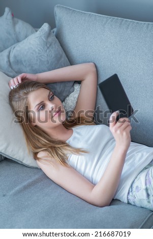 Attractive young woman relaxing on a sofa at home reading an e-book or information on her tablet