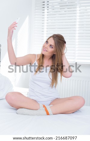 Young woman posing for a selfie on her mobile sitting cross legged on her bed in her sleepwear smiling up at the camera and mussing her hair seductively with her hand