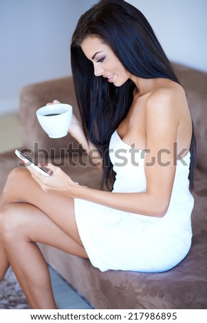 Young woman sitting texting a message on her mobile phone as she sits wrapped in a towel on a sofa drinking coffee