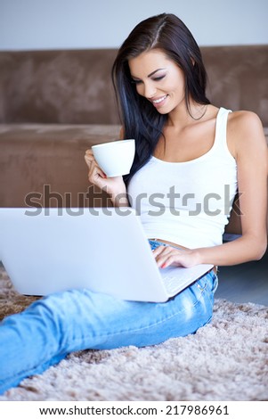 Woman drinking coffee as she types on her laptop computer while relaxing sitting on the carpet leaning against a sofa